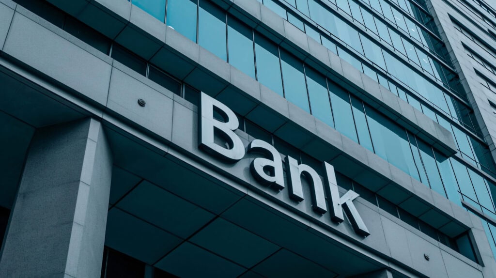 Das Bild zeigt die Fassade eines modernen Bürogebäudes aus Glas und Beton. Im Vordergrund ist ein großes Schild mit dem Wort „Bank“ in weißen, dreidimensionalen Großbuchstaben zu sehen. Die Glasfenster des Gebäudes spiegeln den Himmel und verlaufen in vertikalen Reihen. Die Aufnahme ist aus einem schrägen, nach oben gerichteten Winkel gemacht, wodurch die Höhe des Gebäudes betont wird.