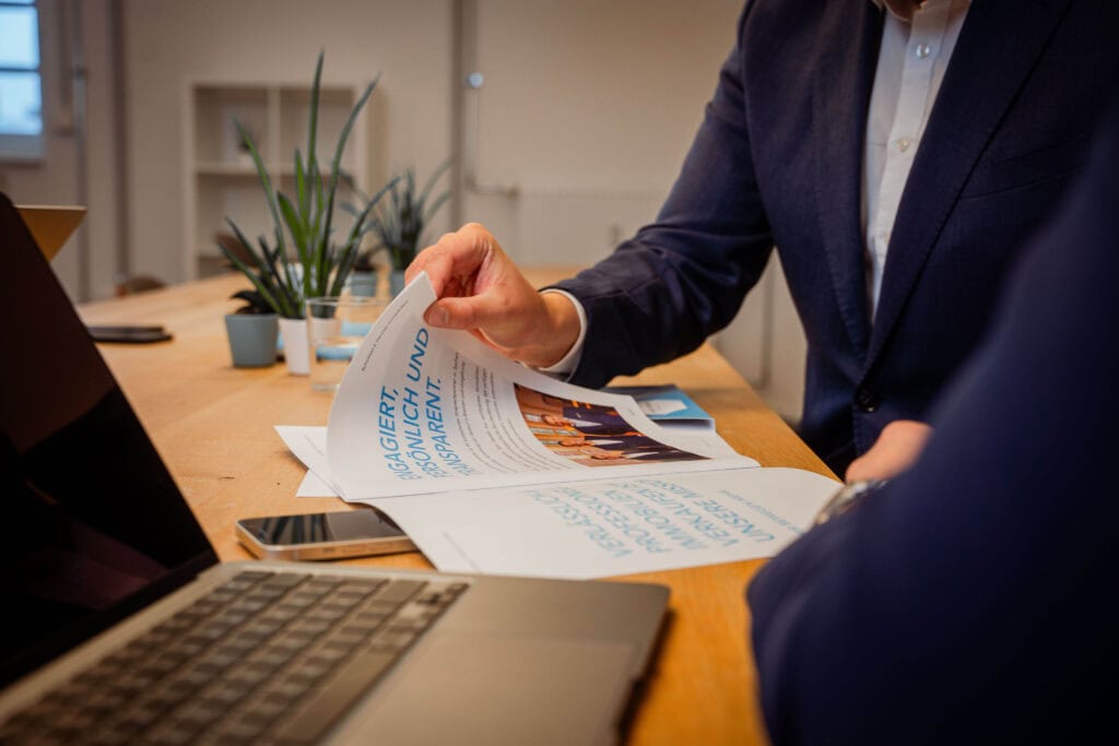 Zwei Personen im Anzug sitzen an einem Holztisch mit Laptop, Smartphone und Broschüren. Eine Person blättert in einer Broschüre mit blauem Text und einem Foto. Im Hintergrund stehen Pflanzen.
